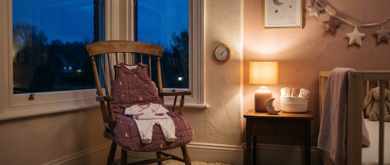 A baby sleep bag and sleepsuit laid out on a wooden chair in a warm lamp-lit nursery