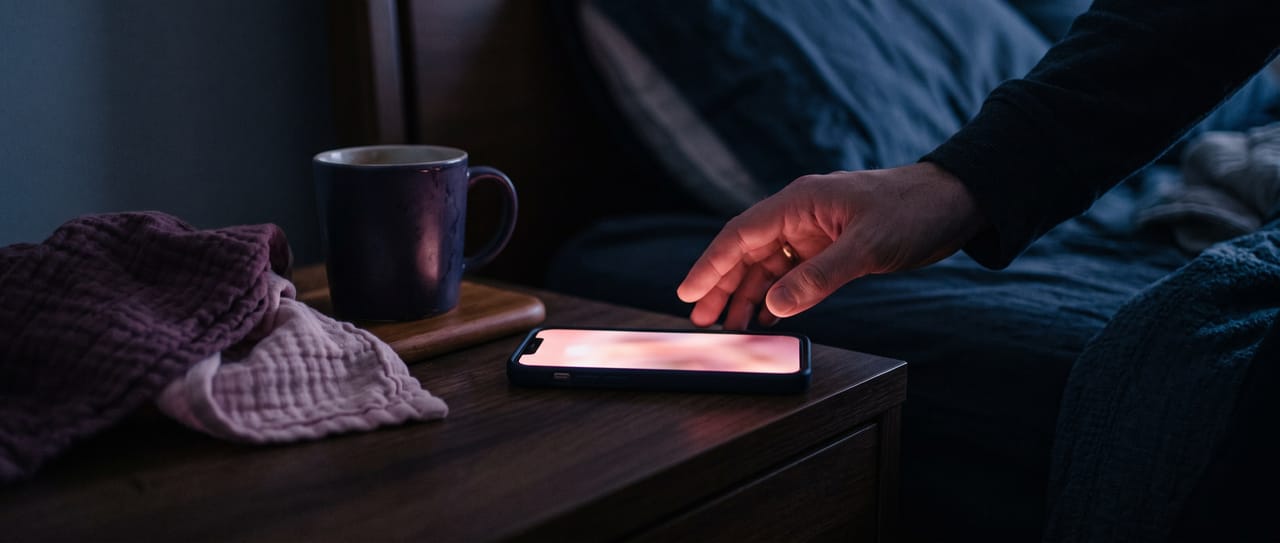 A parent's hand reaching for a glowing phone on a bedside table in the middle of the night