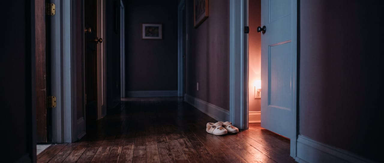 A dark upstairs hallway at night with warm light spilling from a child's bedroom doorway, small slippers by the door