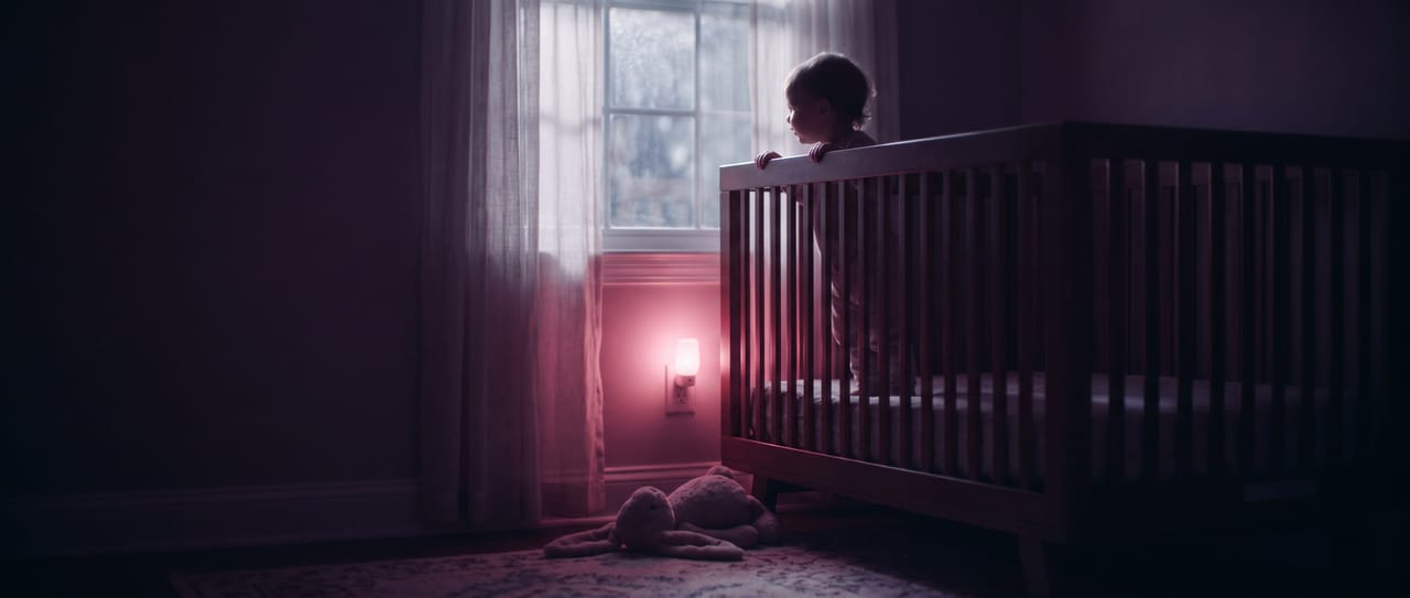 A small child standing up in their cot silhouetted against the window in a dusky, rose-lit nursery